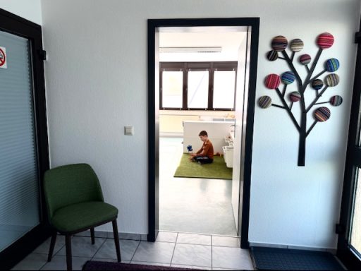 Im Vordergrund ist der Wartebereich mit einem Stuhl und einer Garderobe, im Hintergrund ein Raum, in dem ein Junge auf dem Boden sitzt.